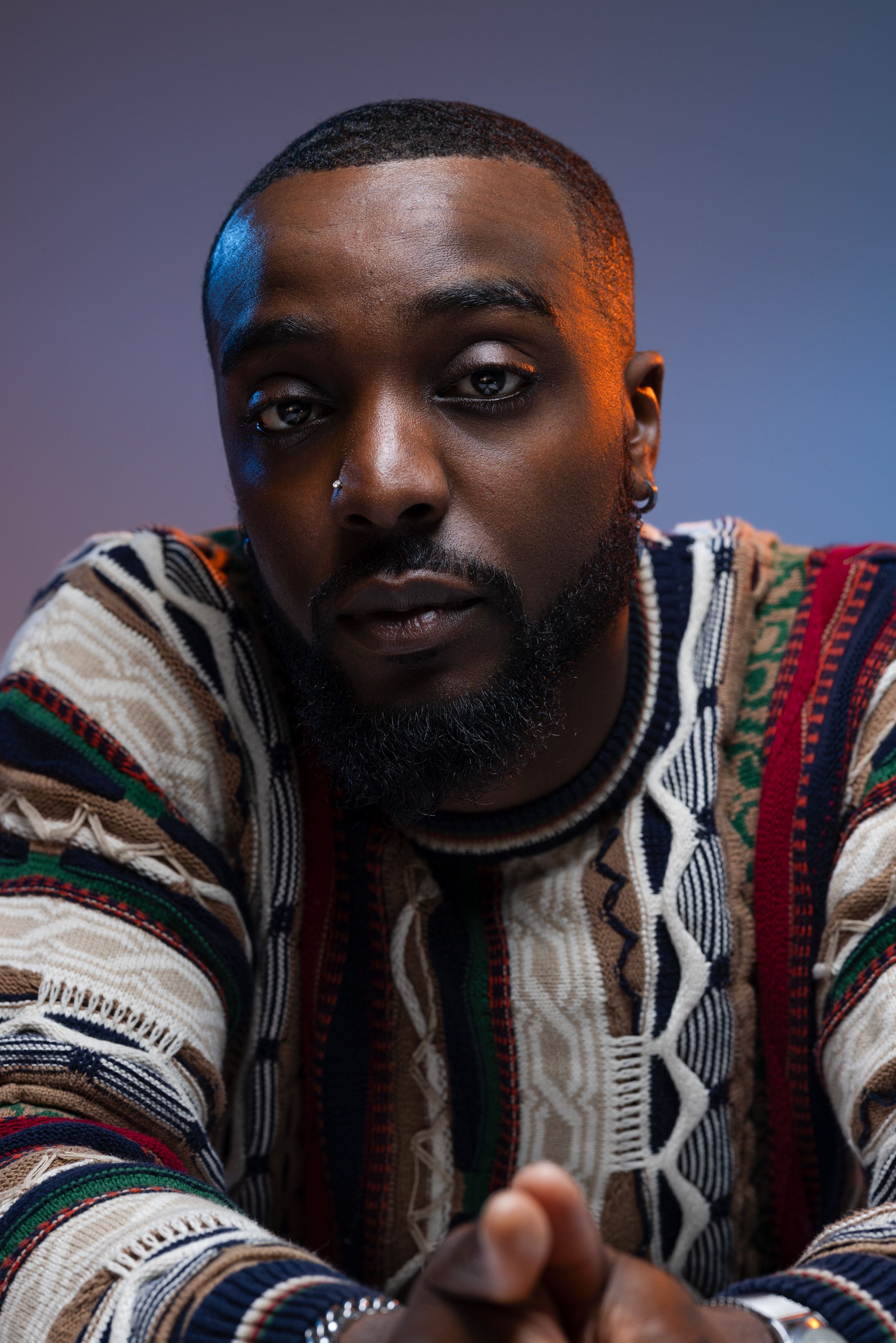 Close-up portrait of someone wearing a patterned knit sweater with colourful stripes against a dark gradient background.