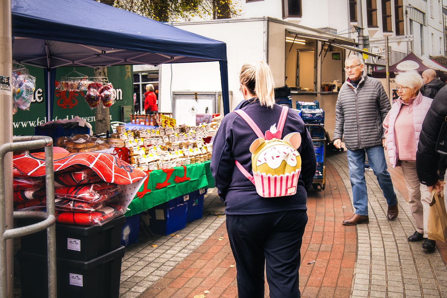 Street market scene featuring a woman with a cupcake-shaped backpack walking past a market stall selling cakes.