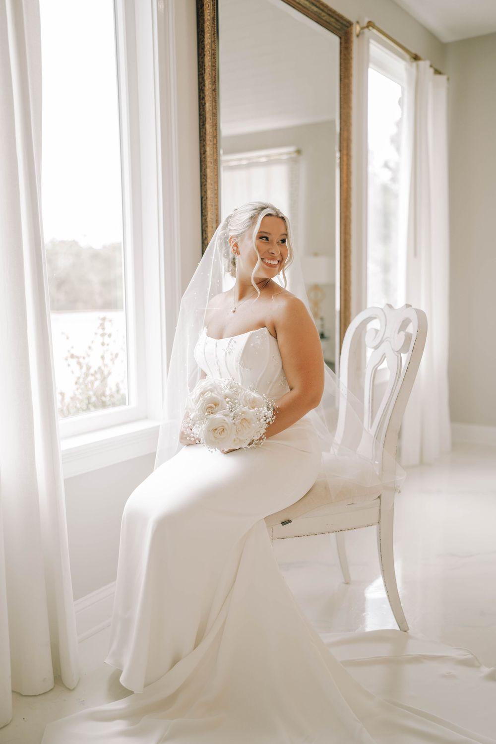 A bride in an elegant white strapless wedding gown sits on an antique chair holding a white bouquet by sunlit windows.