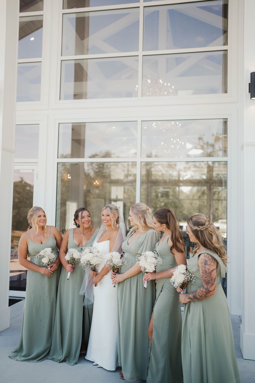 Wedding party in sage green bridesmaid dresses and white bridal gown pose together in front of glass windows.
