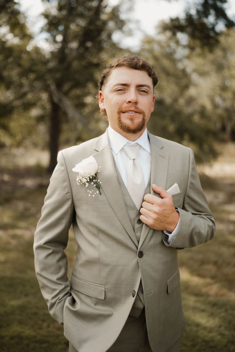 A groom in a light gray suit and white tie adjusts his boutonniere while standing outdoors in a natural setting.