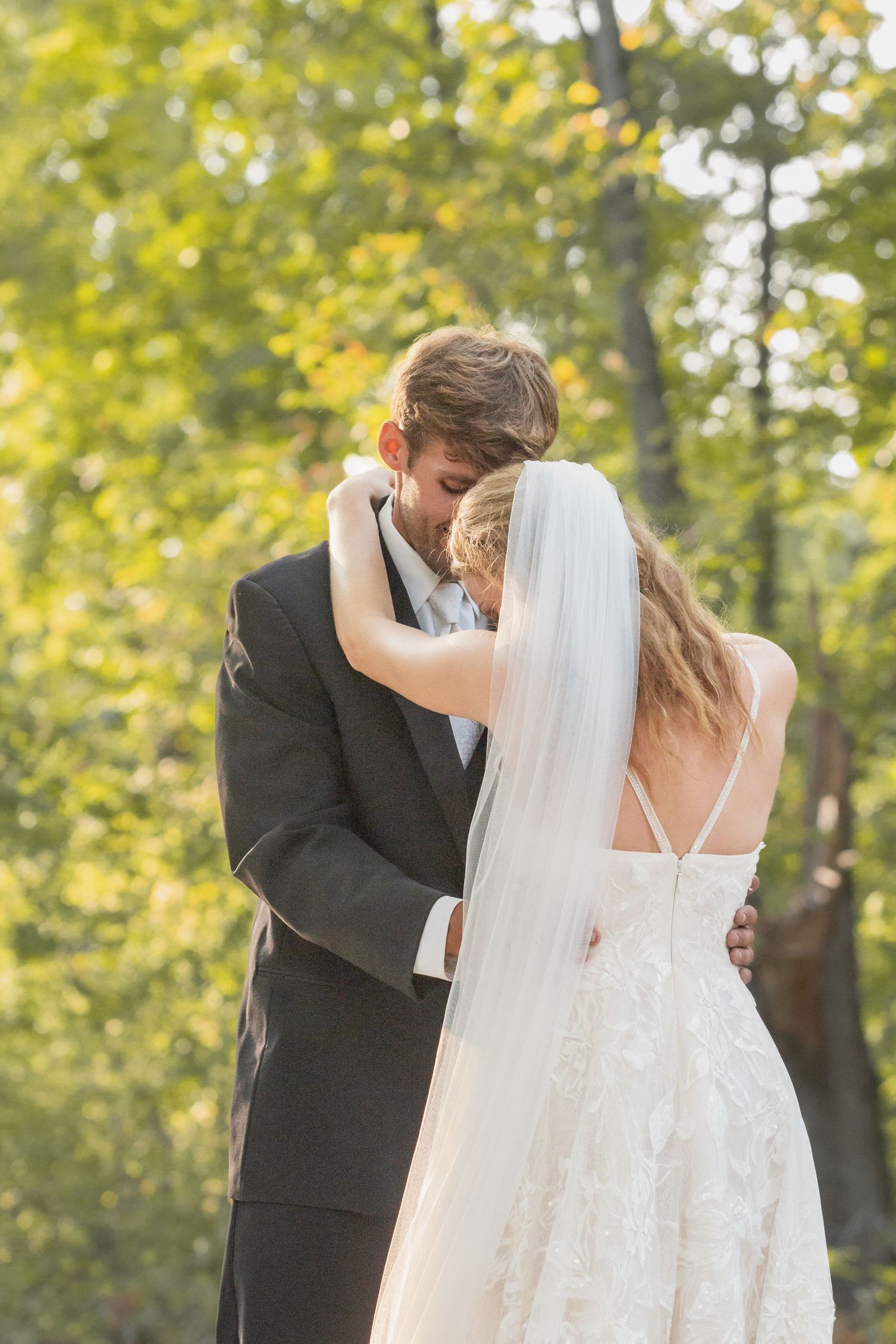 A romantic wedding couple embraces in an outdoor setting with sunlit trees and autumn foliage in the background.