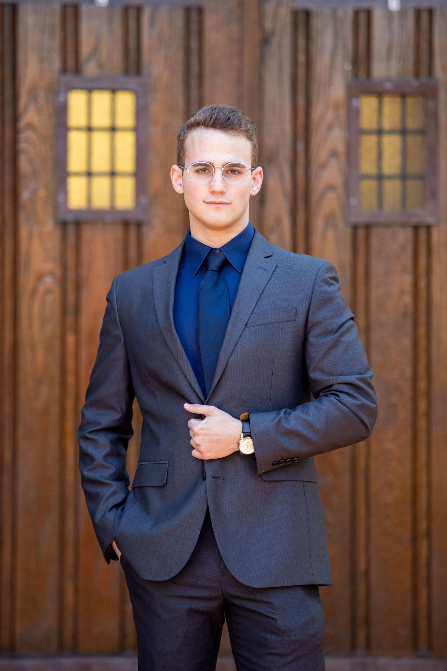 Confident young man in a gray suit and blue tie posing in front of a rustic wooden barn door.