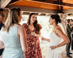 Wedding guests in white and floral dresses mingle and chat at an indoor reception.