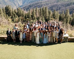 Large wedding party group photo taken outdoors against a backdrop of mountain scenery and evergreen trees.