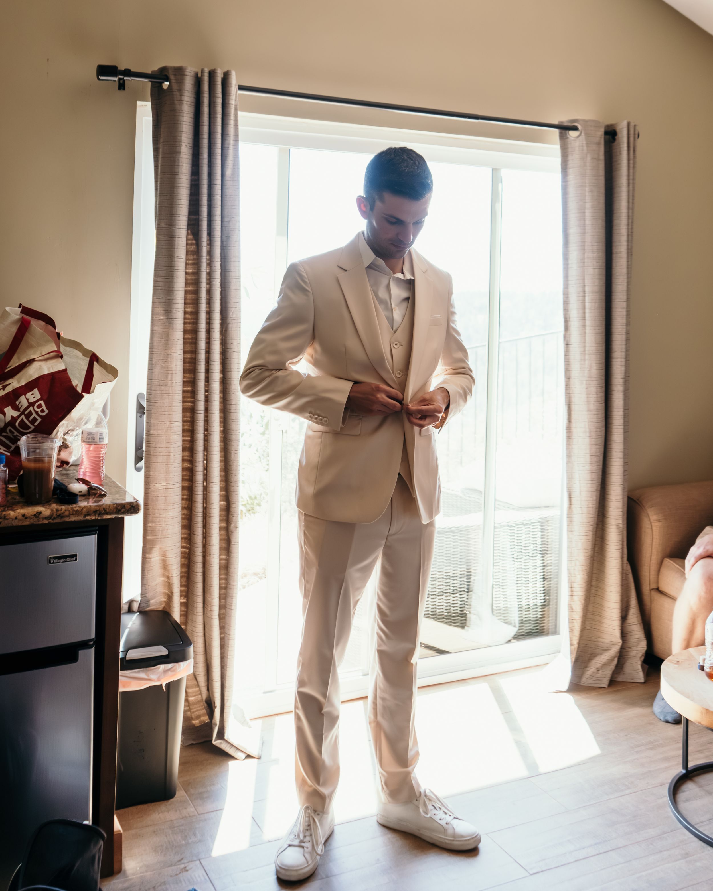 Person in light colored formal attire getting ready in a sunlit room with curtained windows.