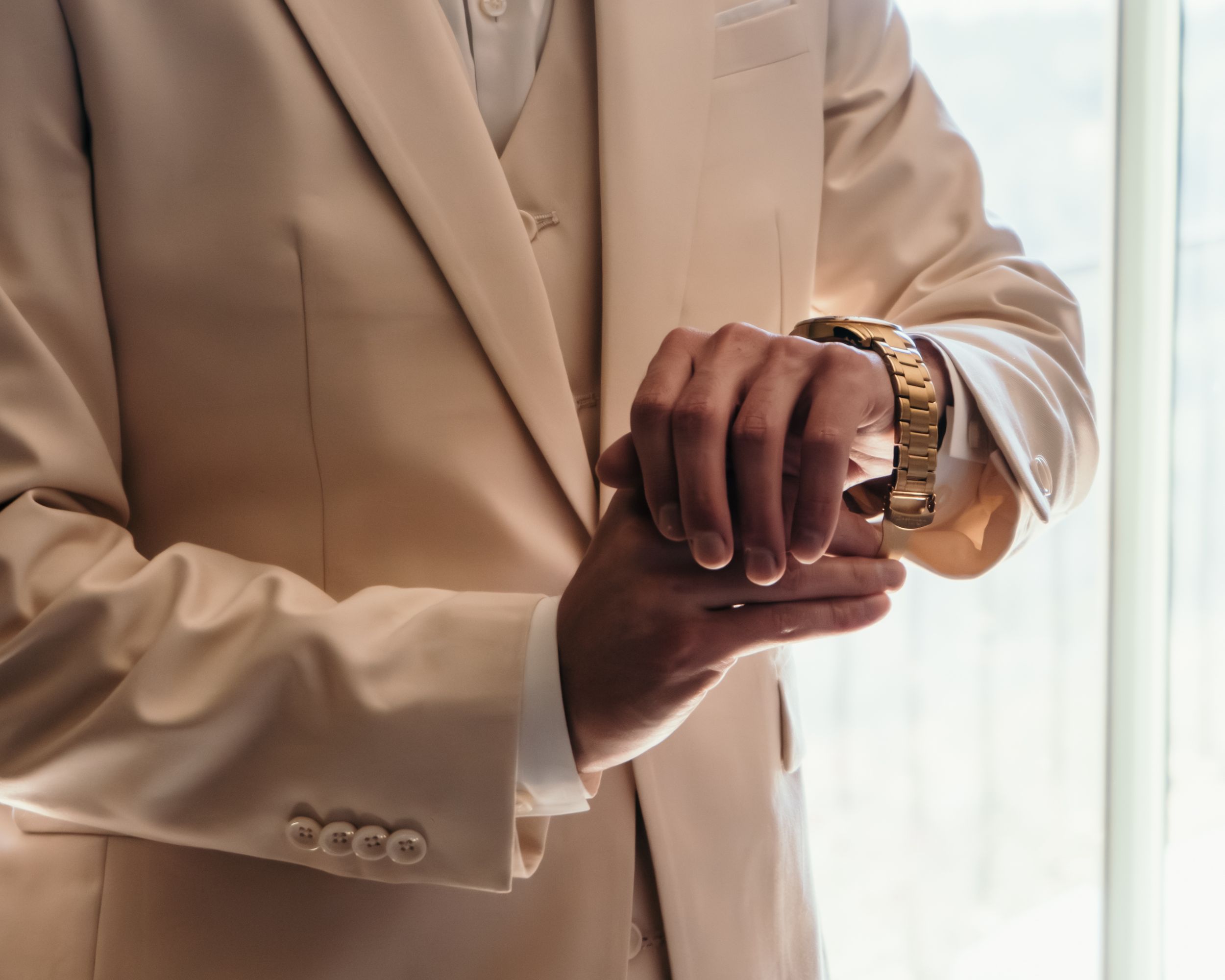A person wearing a beige suit jacket adjusts their sleeve cuff while standing near a window.