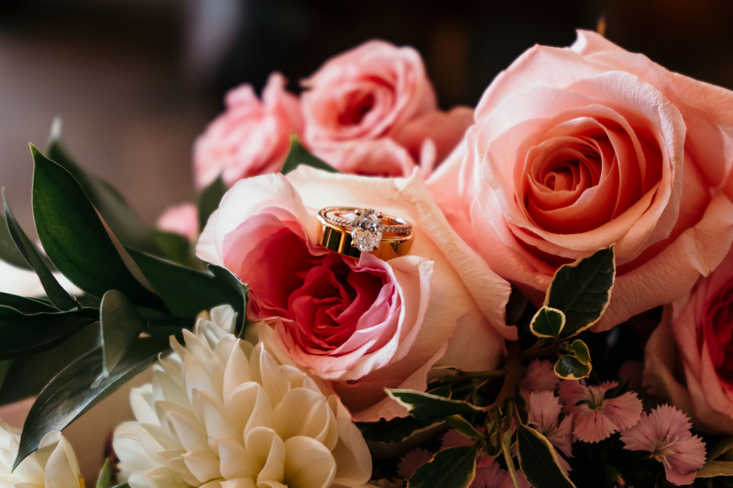 Wedding rings nestled among pink roses and white dahlias in an elegant floral arrangement.