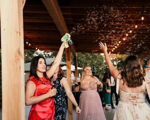 Wedding guests dancing with raised arms under a wooden pavilion decorated with string lights.