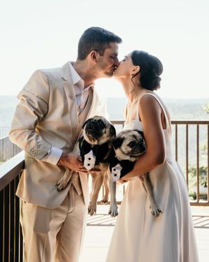 Newlyweds in white and beige attire sharing sweet moments with their pug dog on a balcony.