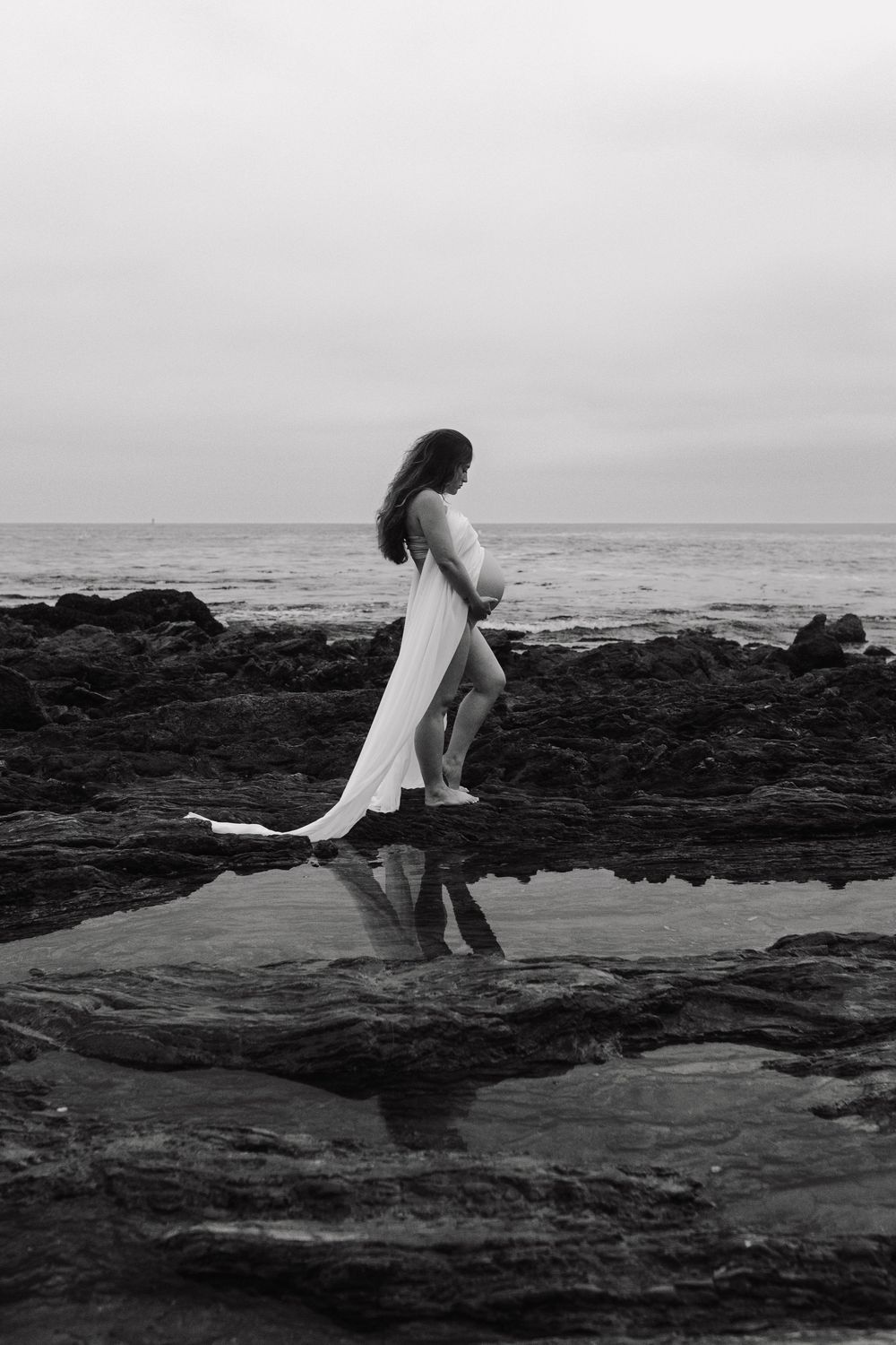 A black and white sequence shows a figure in white dress moving gracefully across rocky coastal terrain at the ocean's edge.