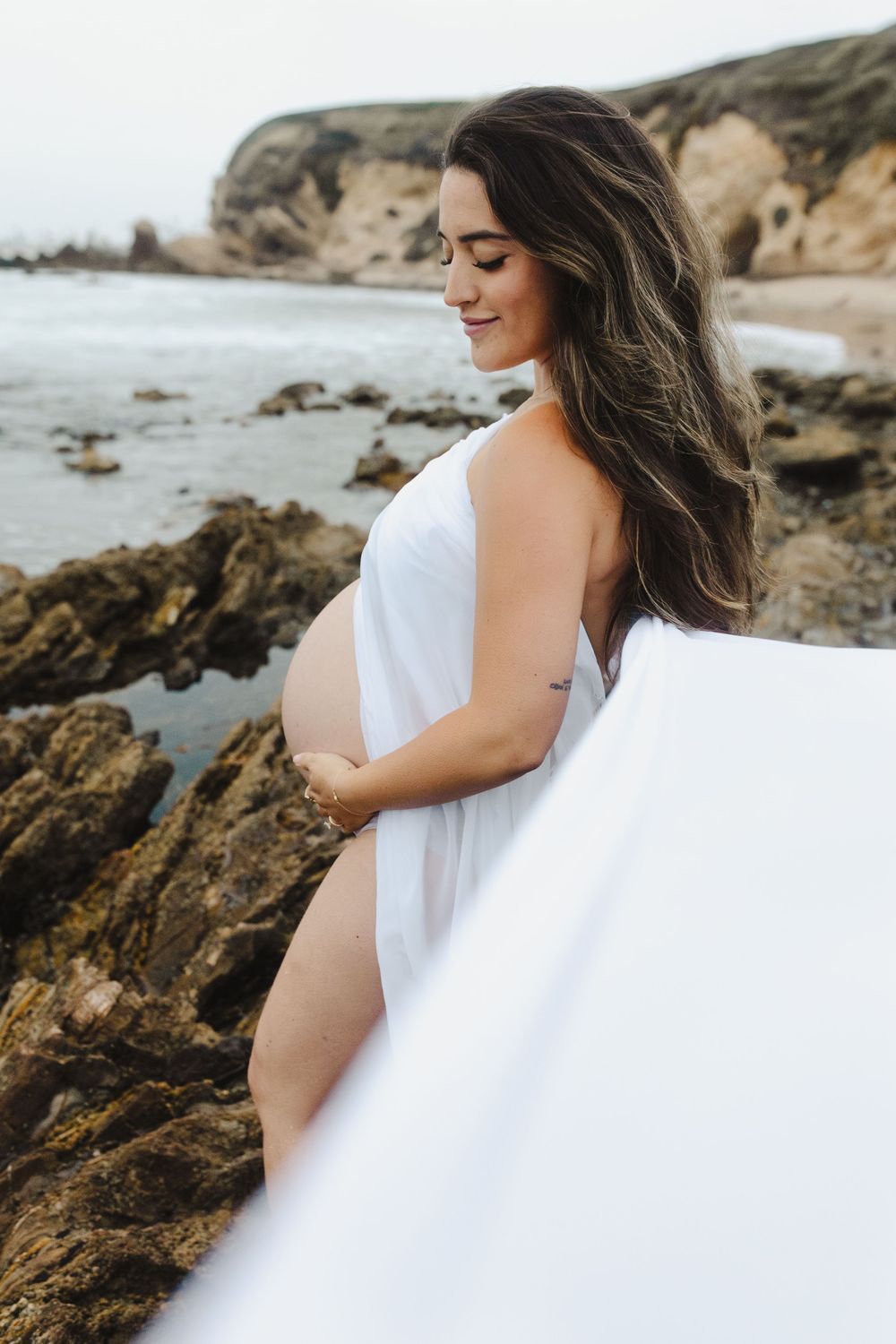 A woman in a flowing white dress cradles her pregnant belly while standing on a rocky beach at sunset.