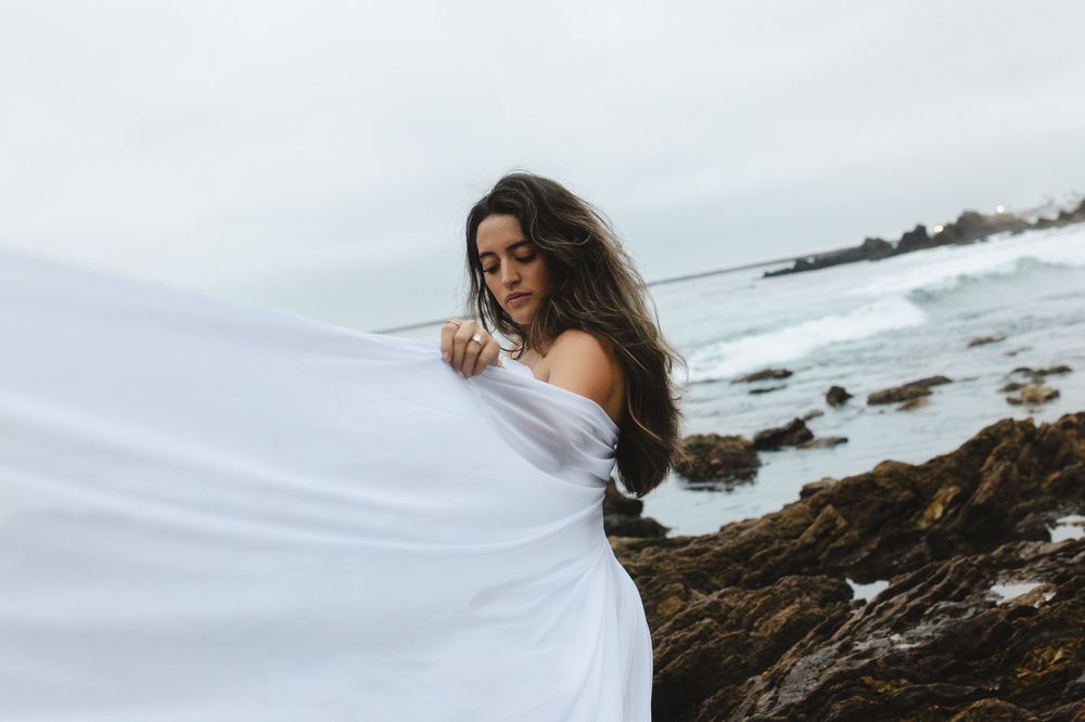 Woman in white dress poses dramatically on rocky coastline with crashing waves in the background.