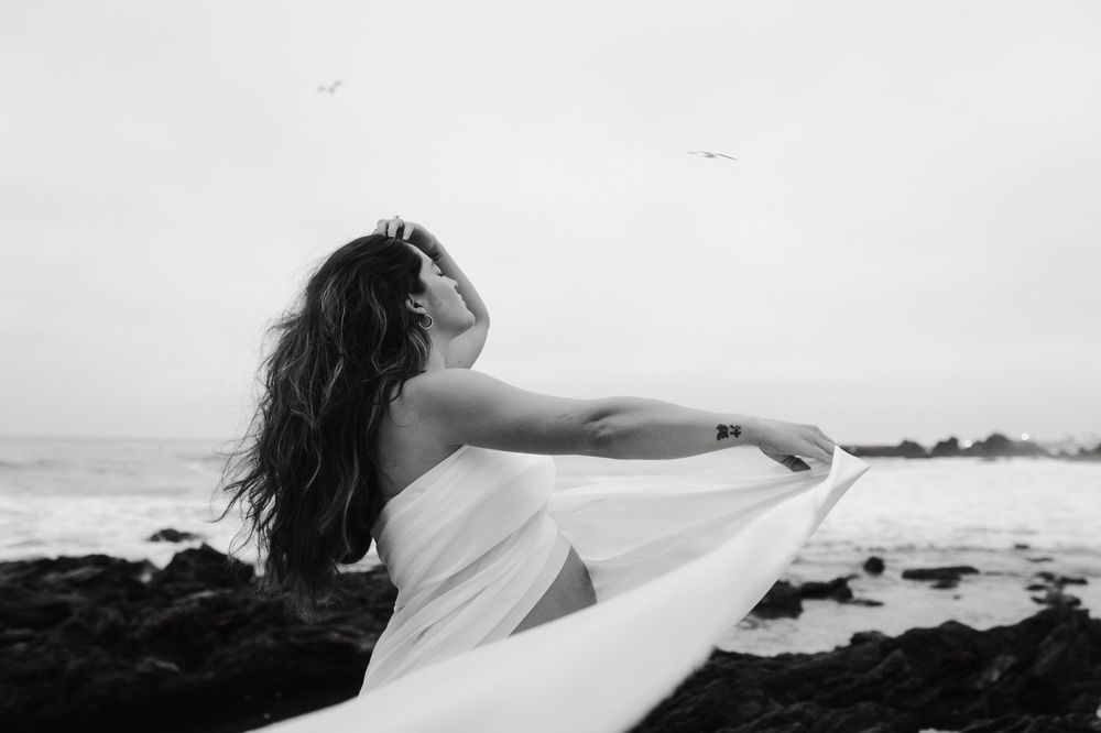 Black and white photo of a figure in a white dress with flowing fabric standing on a rocky beach by the ocean.