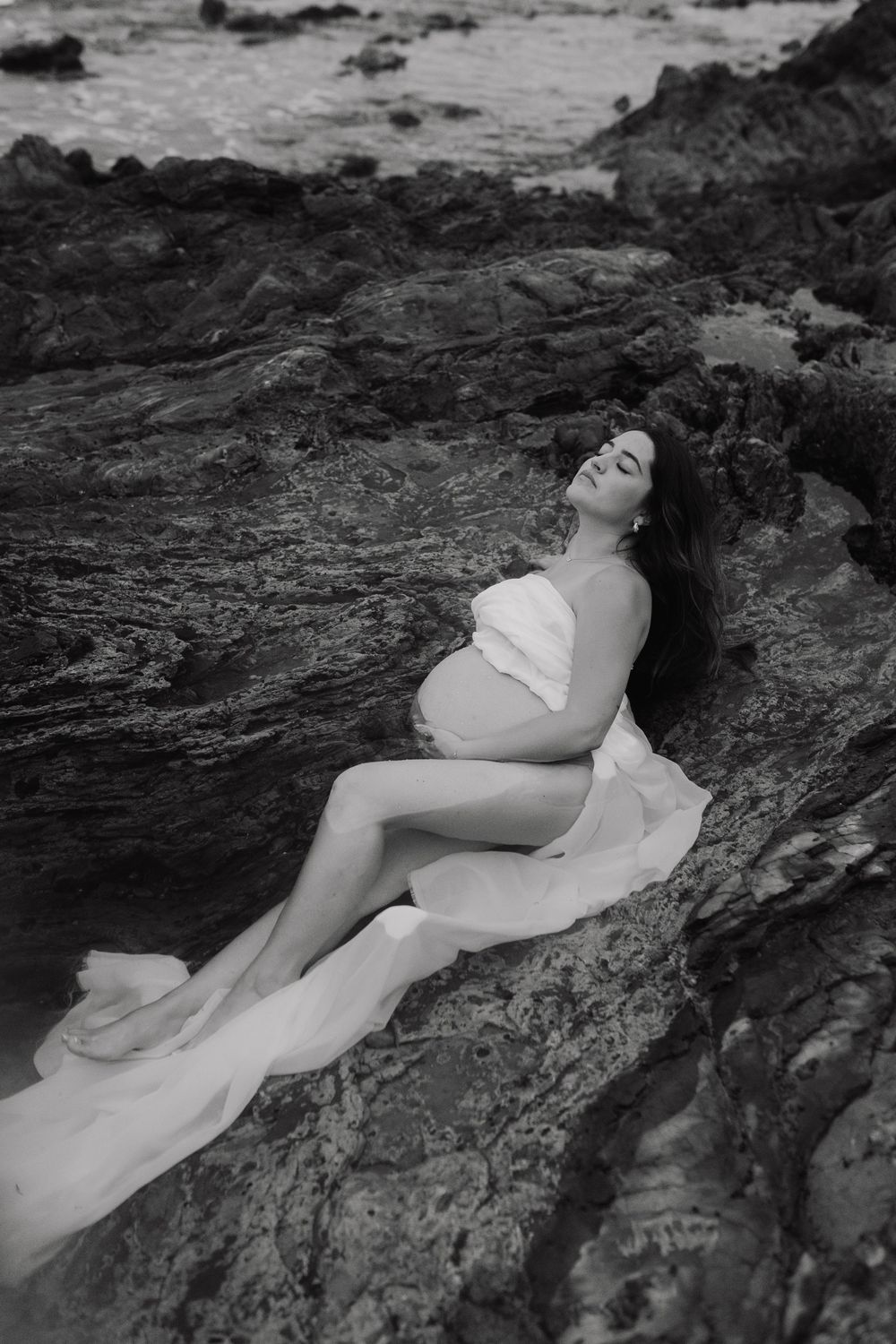A figure in a white dress reclines dramatically across dark rocky terrain in a black and white artistic photograph.