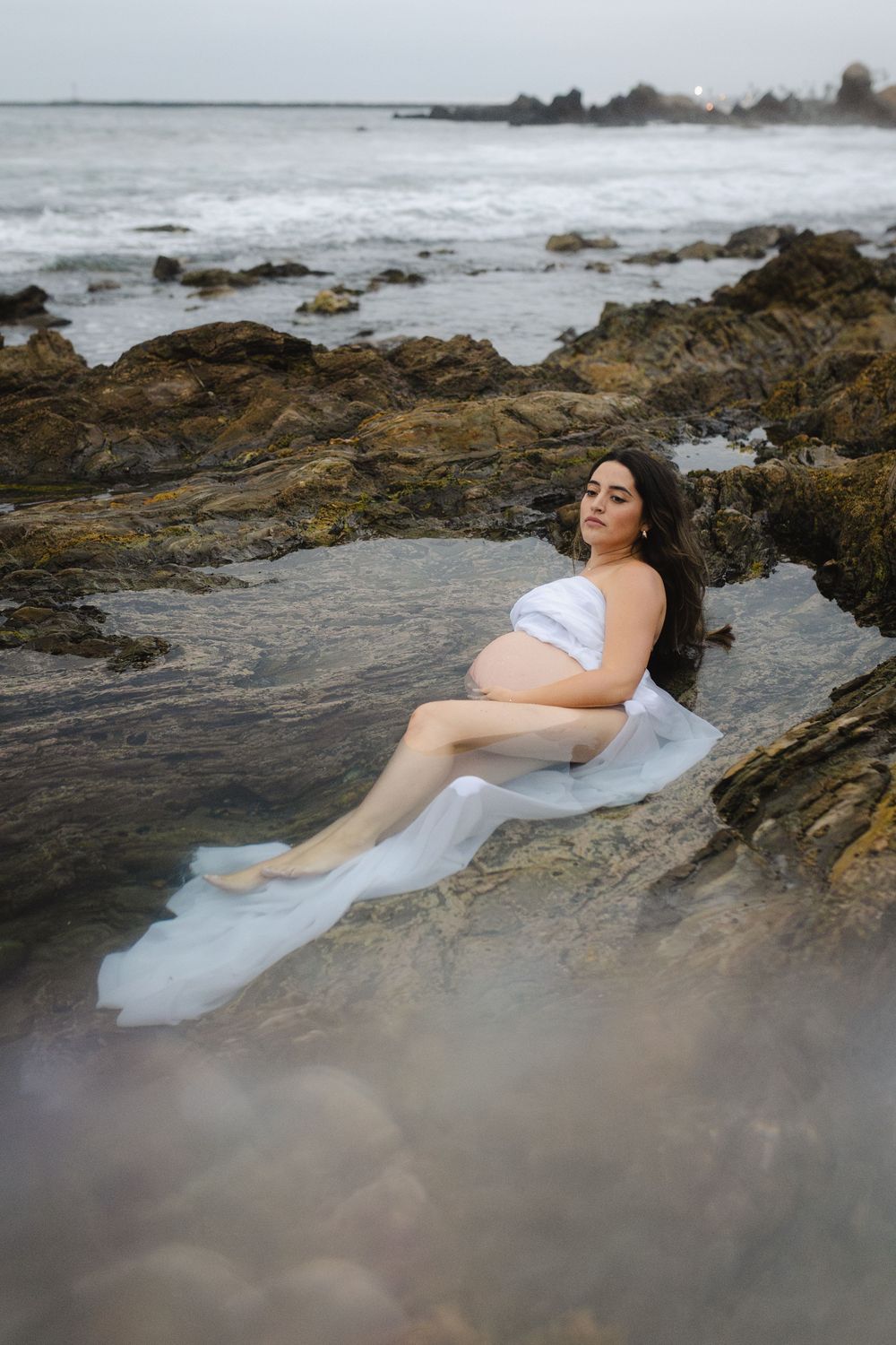 Ethereal coastal scene with flowing white fabric draped over rocky shoreline as waves crash in background.