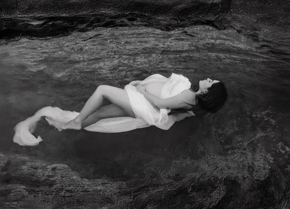 Seven black and white photographs showing a figure in white flowing fabric laying on dark rocky terrain.