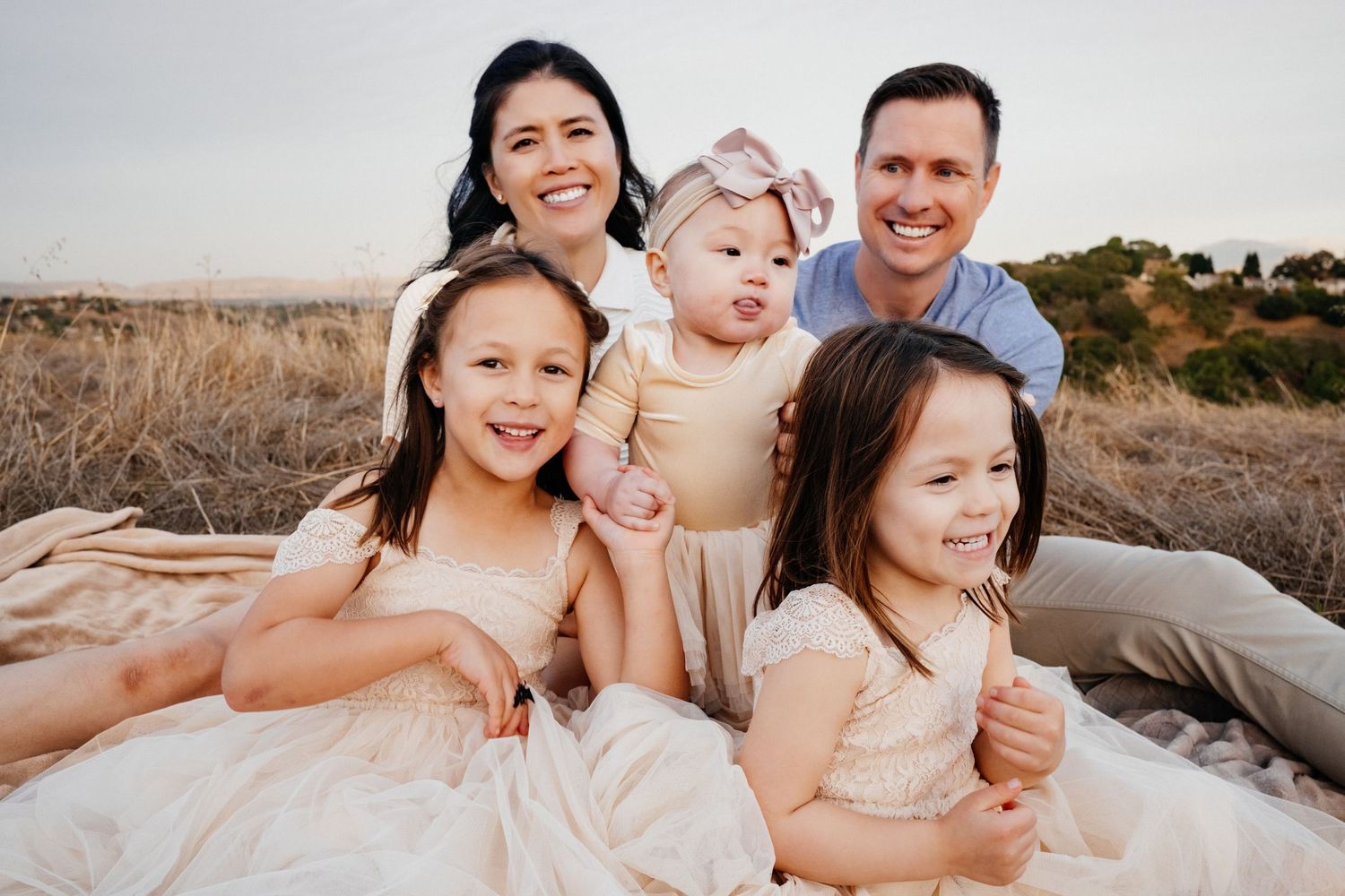 A joyful family wearing neutral colors poses together outdoors during a sunset photo session at the beach.