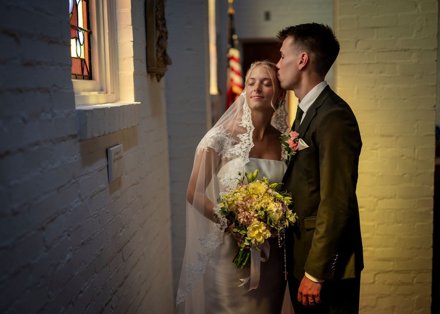 A romantic wedding couple shares an intimate moment in soft window light with a yellow bouquet.