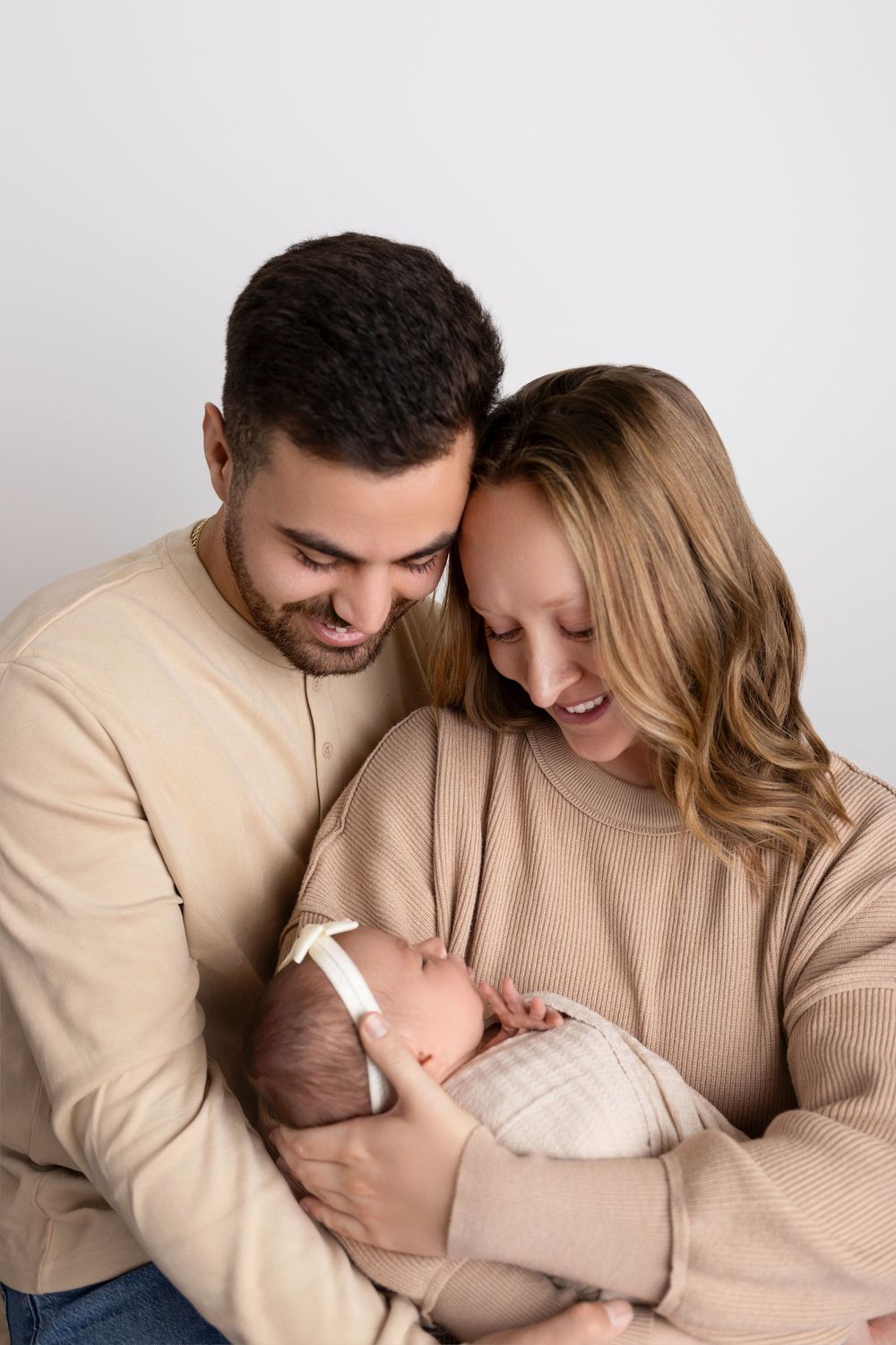 A couple in matching beige sweaters gazes lovingly at their newborn baby against a light background. NJ newborn photographer in Sussex Count