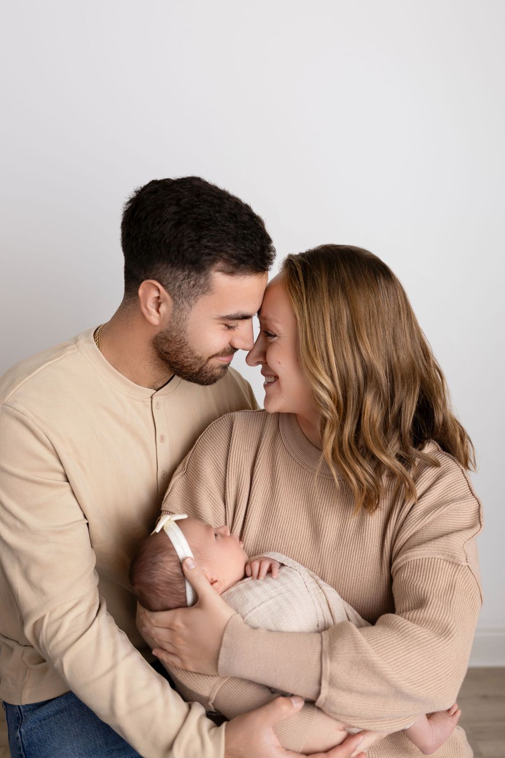 New parents in beige sweaters share an intimate moment while holding their newborn baby against neutral background. NJ newborn photographer