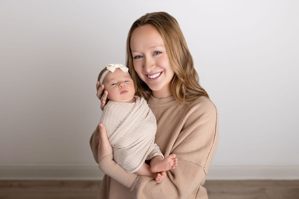 A person in a beige sweater holds a swaddled newborn baby against a neutral gray wall. NJ newborn photographer in Sussex County