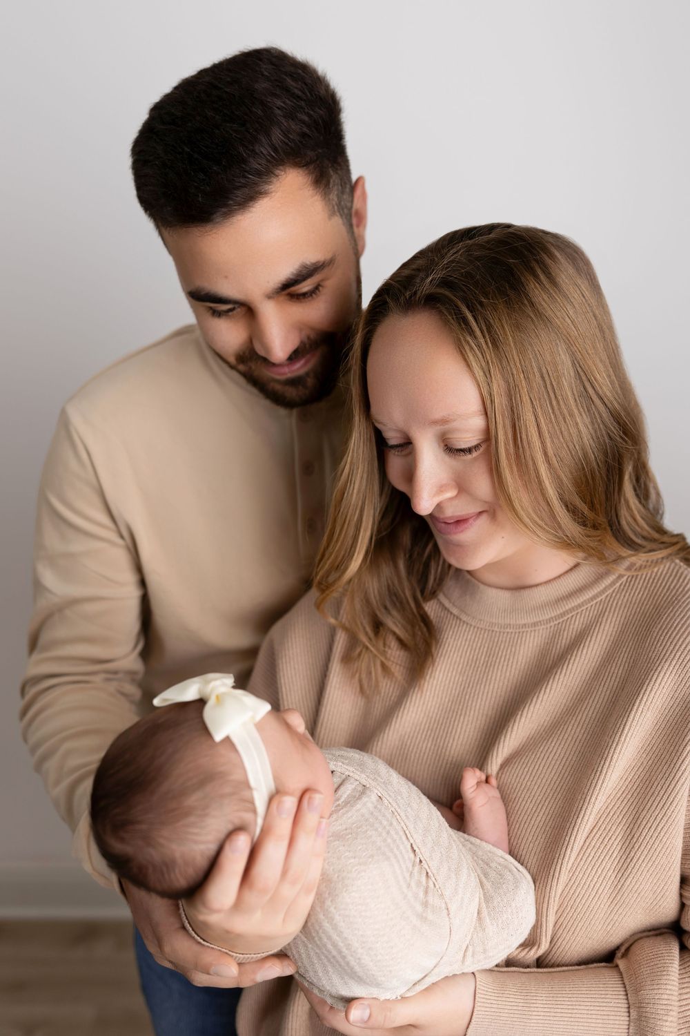 Parents in neutral-colored sweaters share an intimate moment while holding their swaddled newborn. NJ newborn photographer in Sussex County