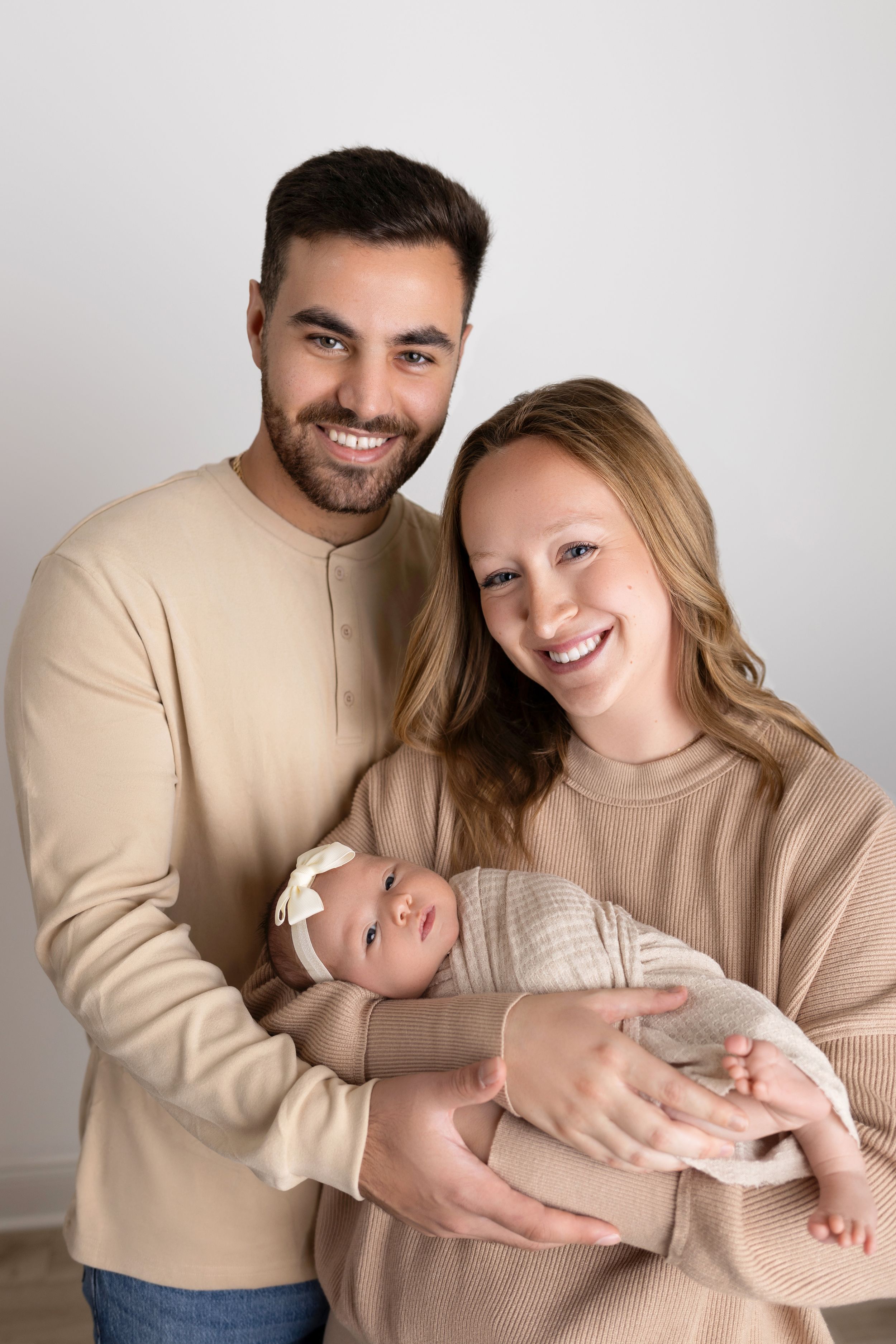 Family of three wearing coordinating beige outfits pose together for a portrait against white background. NJ newborn photographer in Sussex