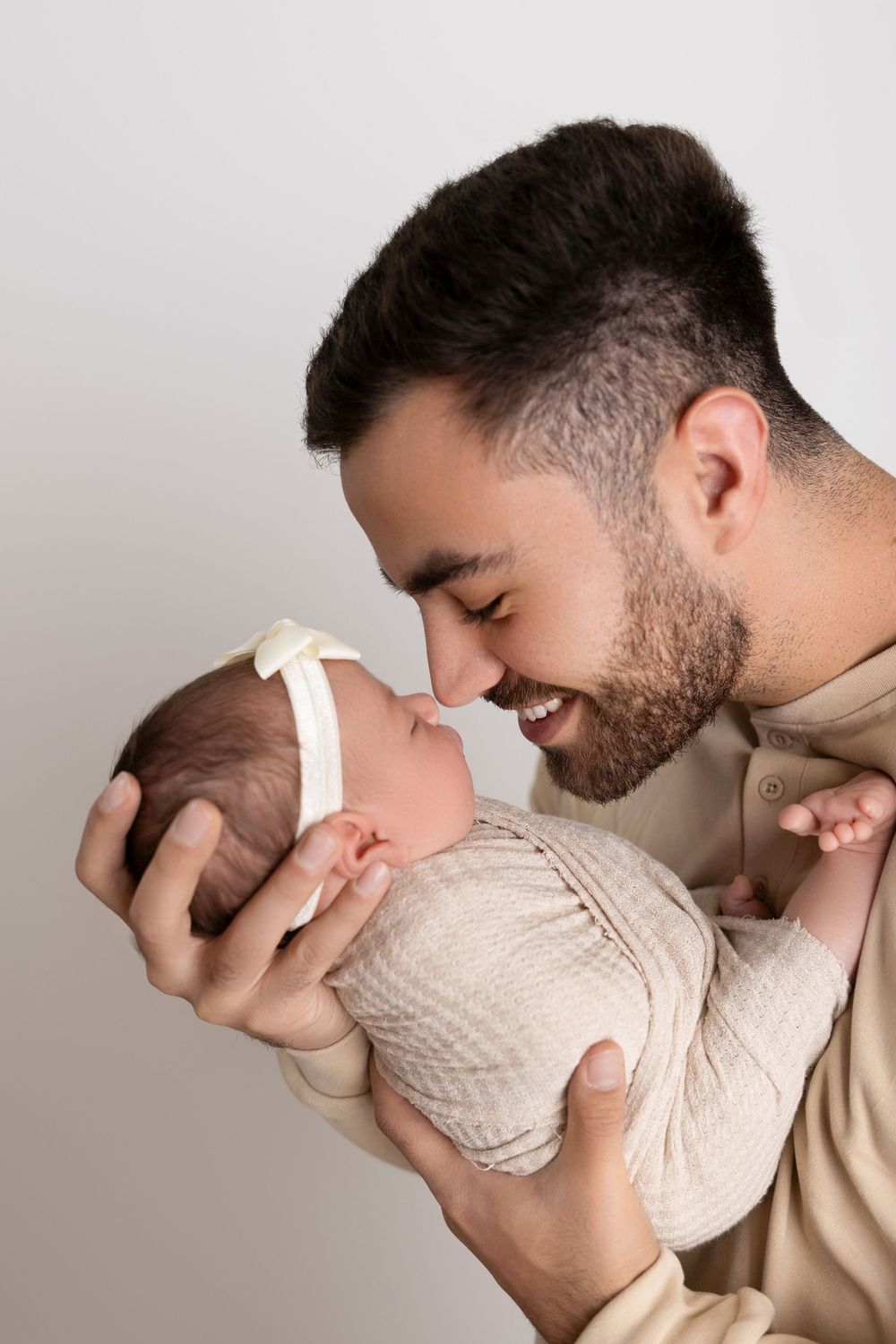 Father in beige sweater shares tender moment with swaddled infant against light background. NJ newborn photographer in Sussex County