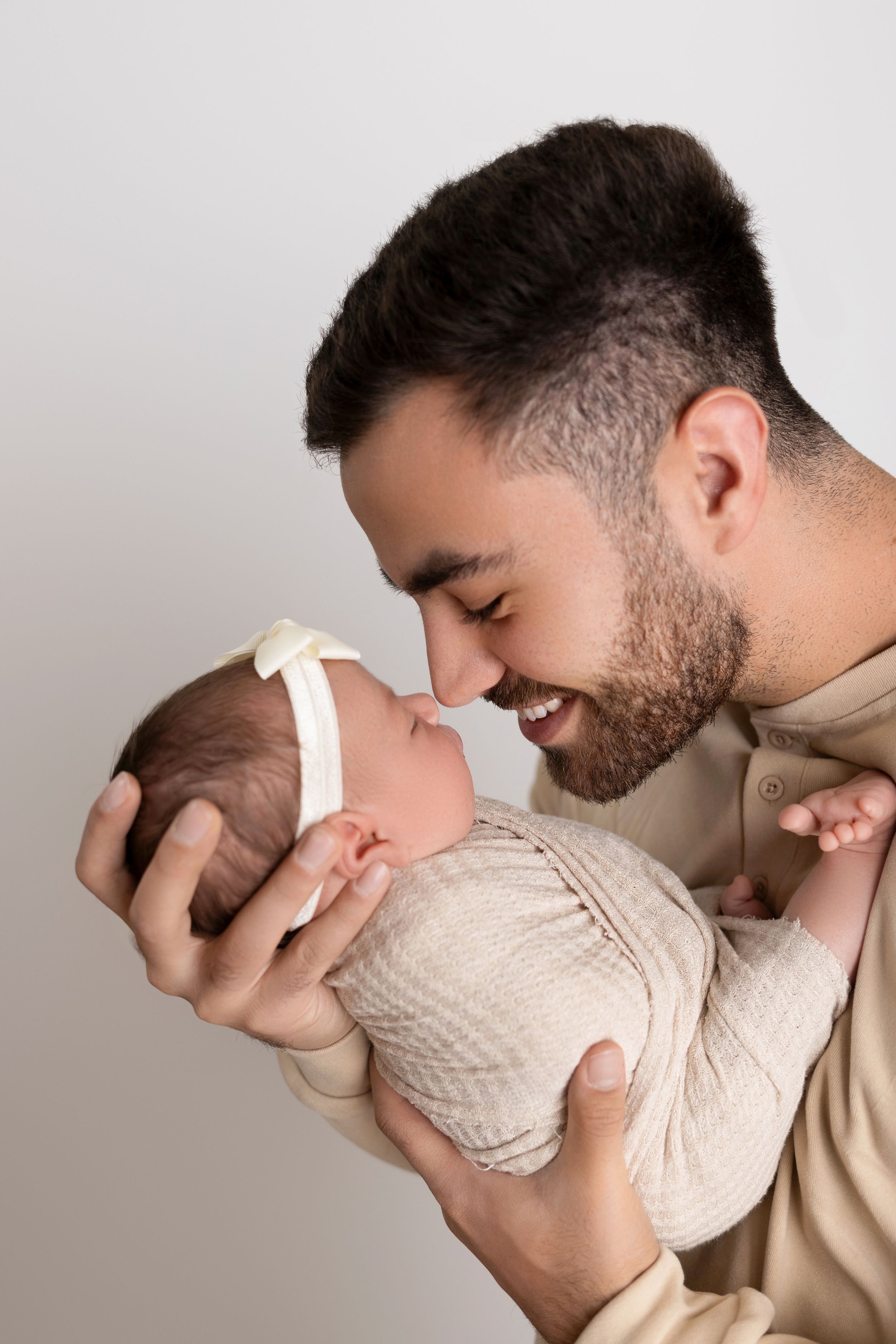 Father in beige sweater shares tender moment with swaddled infant against light background. NJ newborn photographer in Sussex County