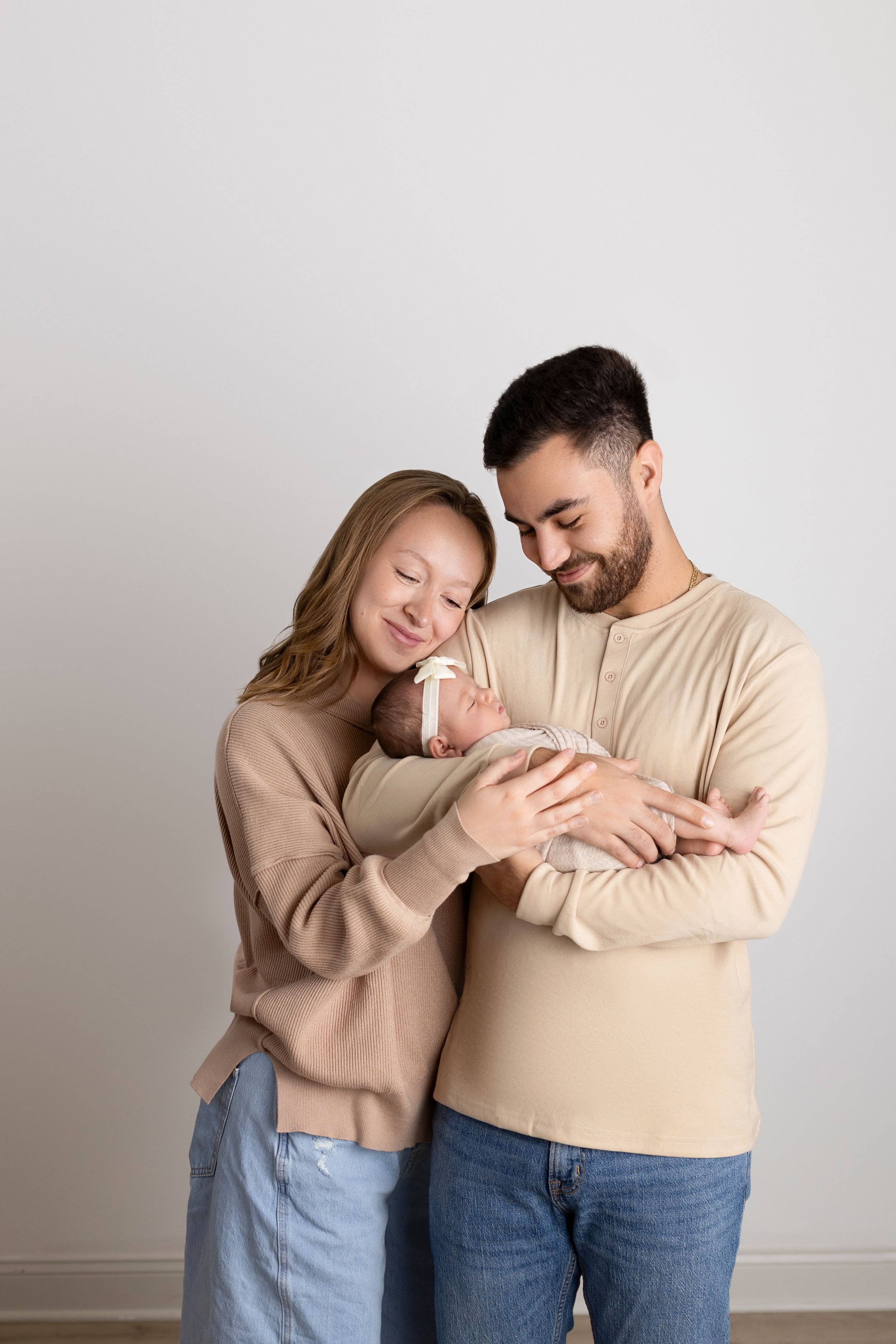 A couple in beige and denim outfits embrace tenderly against a light gray background. NJ newborn photographer in Sussex County