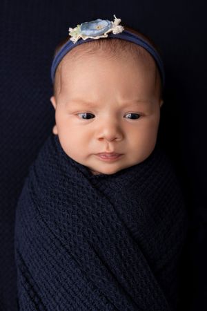 Baby wearing blue floral headband wrapped in navy knit blanket against dark background. NJ newborn photographer in Sussex County