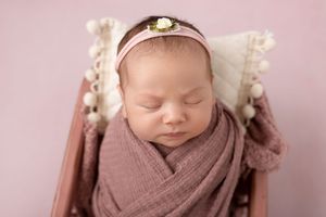 Newborn baby sleeping peacefully wrapped in mauve swaddle with floral headband against pink background. NJ newborn photographer in Sparta NJ