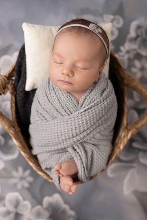 Sleeping newborn baby wrapped in gray knit blanket rests in woven basket with floral backdrop. Sparta NJ photographer