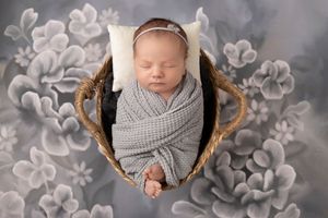 Newborn baby wrapped in cozy gray knit blanket sleeping peacefully in wooden basket with floral backdrop.