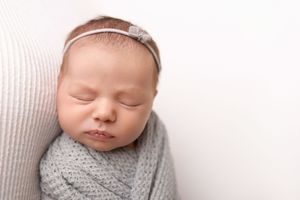 Sleeping newborn baby wearing a gray knit wrap and white headband rests peacefully during professional photo session.