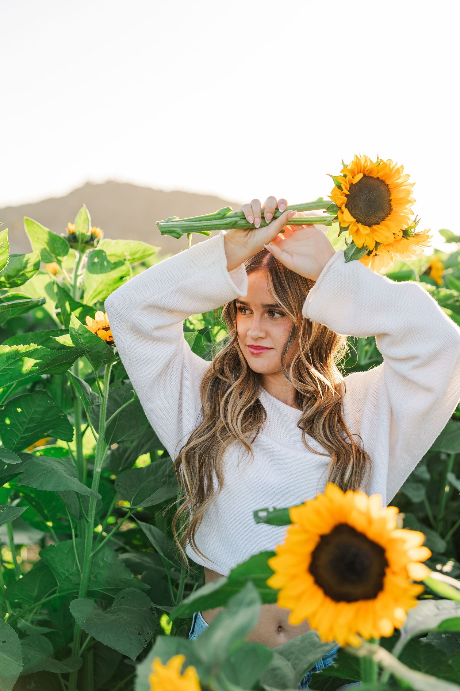 A person in a white sweater poses playfully among bright yellow sunflowers in a garden setting.