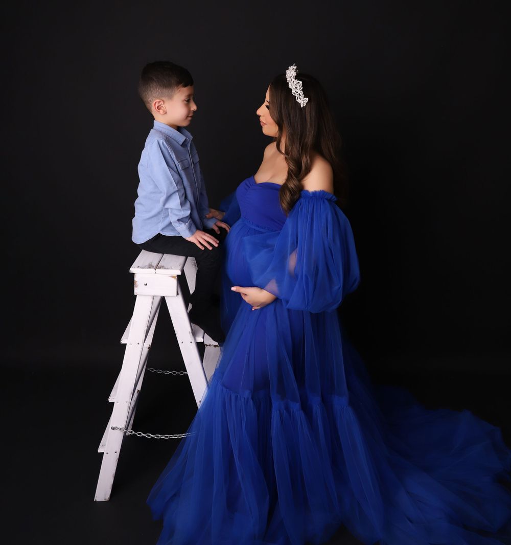 A maternity photo with a child standing on a ladder next to someone in a flowing blue gown.