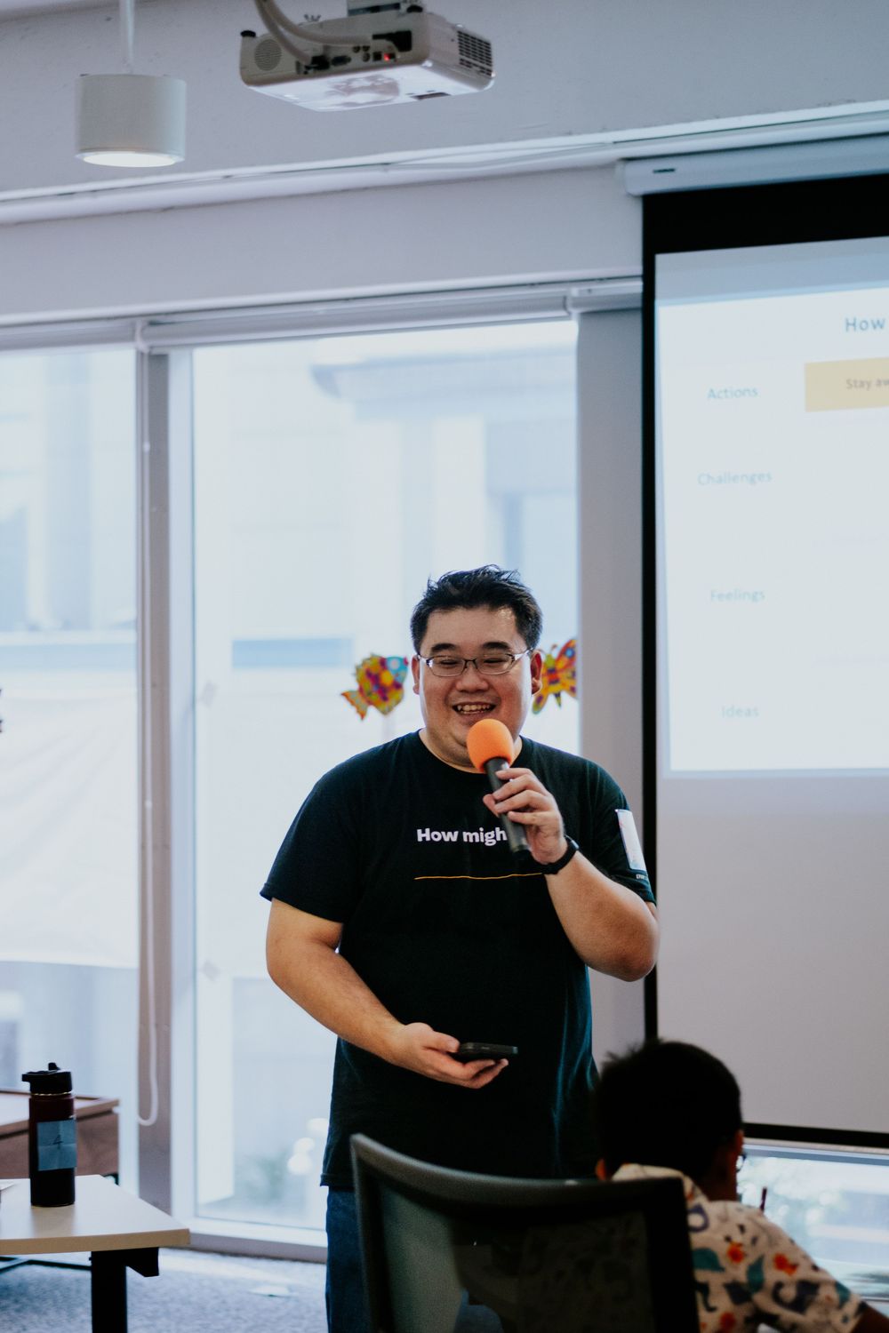 A person in a black shirt sits at a desk in a modern office with large windows and a projector screen.