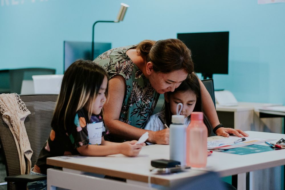 Students working together at a desk in a classroom with turquoise walls and computer monitors in the background.