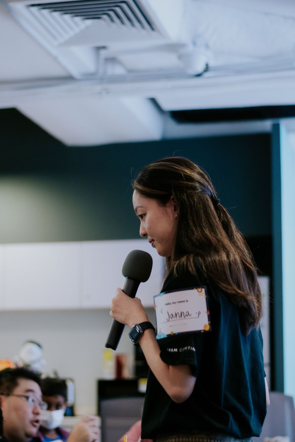 A person in a dark shirt drinks from a coffee cup while speaking into a microphone during a presentation.