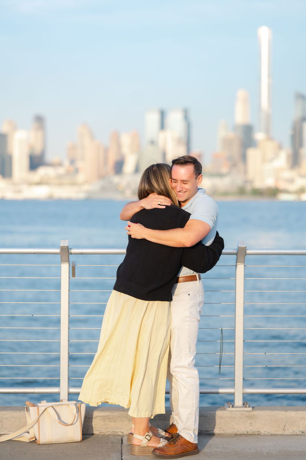 A couple shares an intimate moment along a waterfront railing with a city skyline visible in the background.