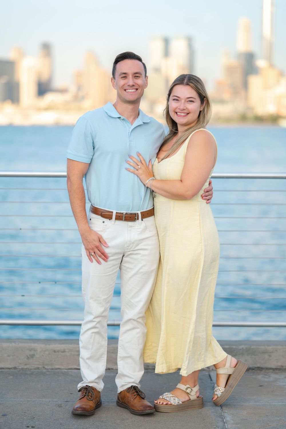 A couple poses together on a waterfront boardwalk with the ocean and city skyline behind them.