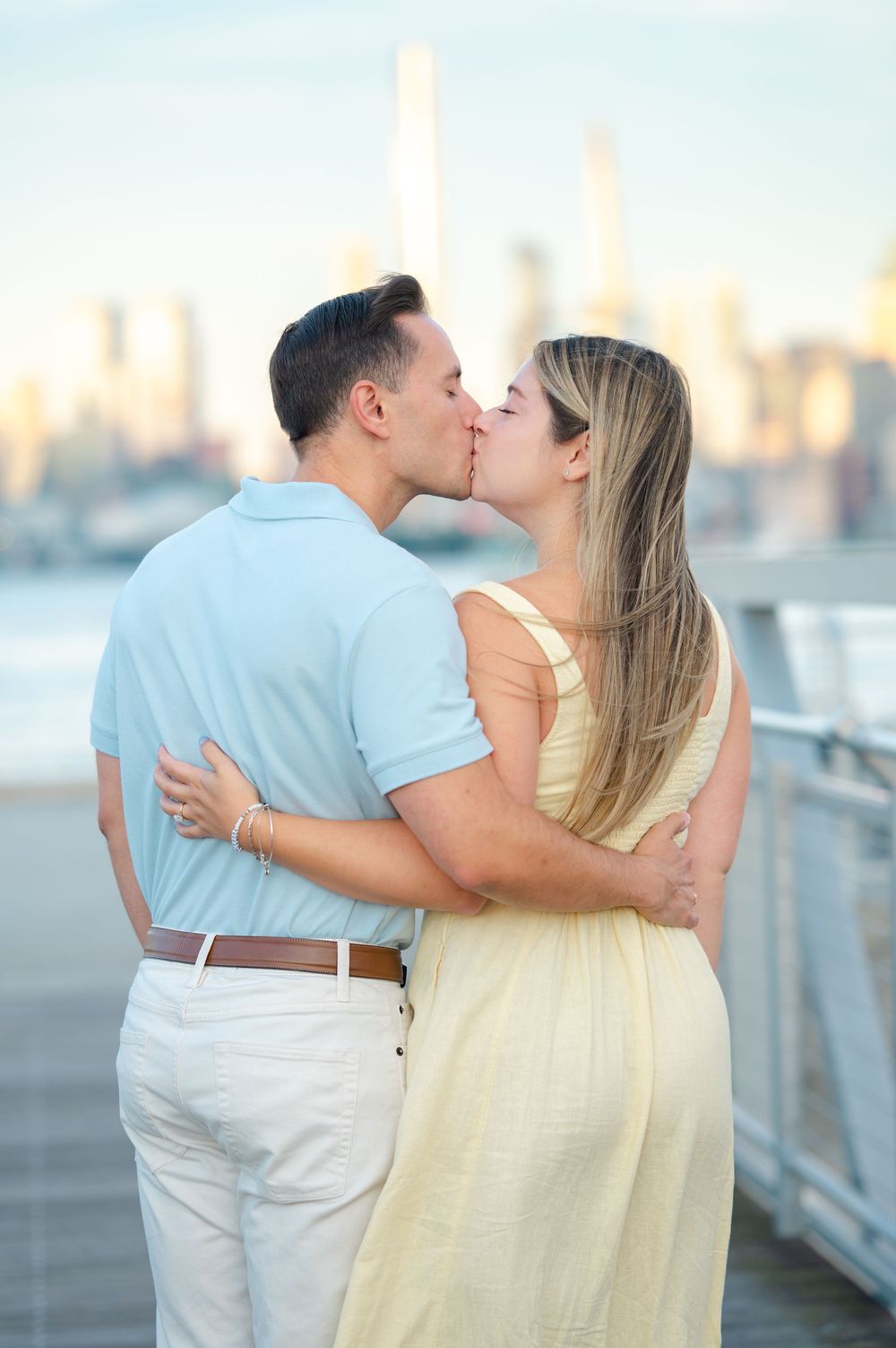 A romantic couple embraces and kisses on the Weehawken Pier with the NYC city skyline visible in the background at sunset.