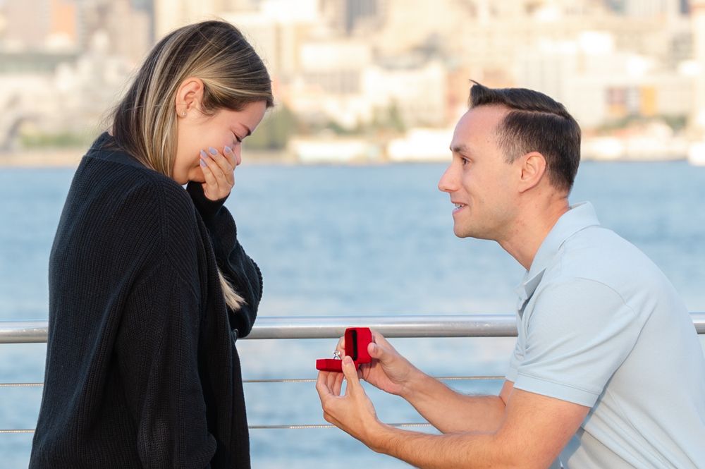 Romantic proposal sequence on a waterfront boardwalk with city skyline in background.