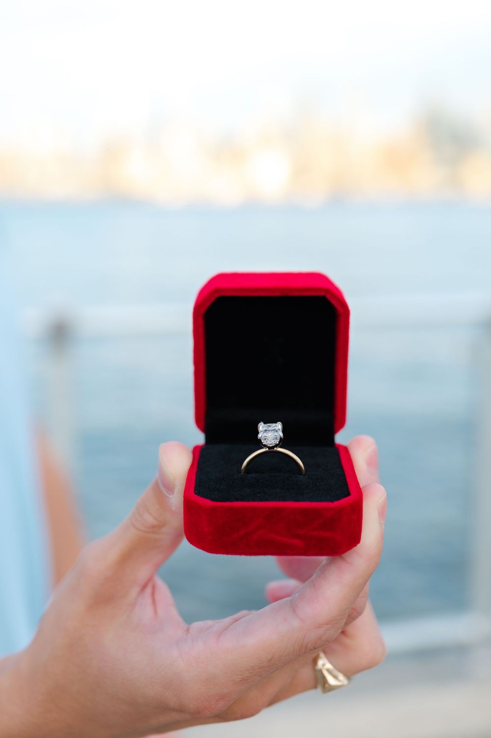 A diamond engagement ring sits in an open red velvet box with a blurred waterfront background.