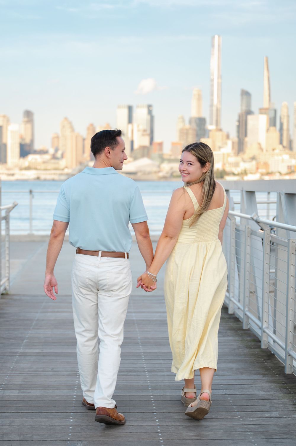 A couple in coordinated pastel outfits walk hand in hand along a waterfront pier with the NYC skyline in the background.