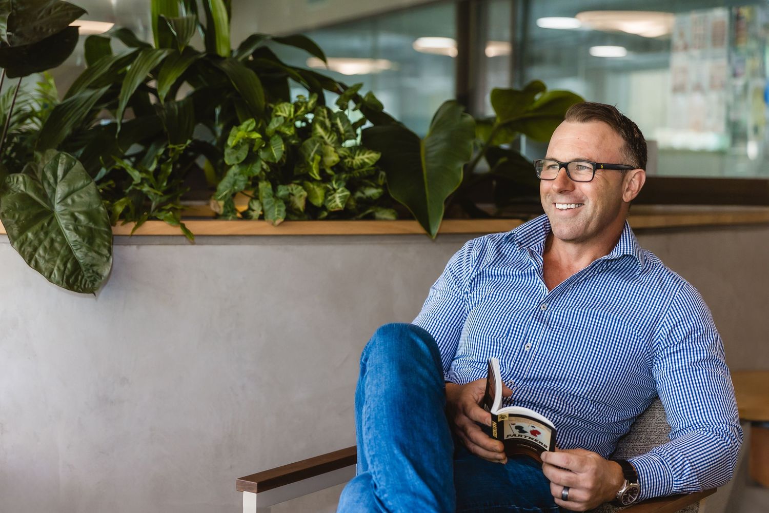 Professional in a blue button-down shirt sits at a table in a modern restaurant with green plants in the background.