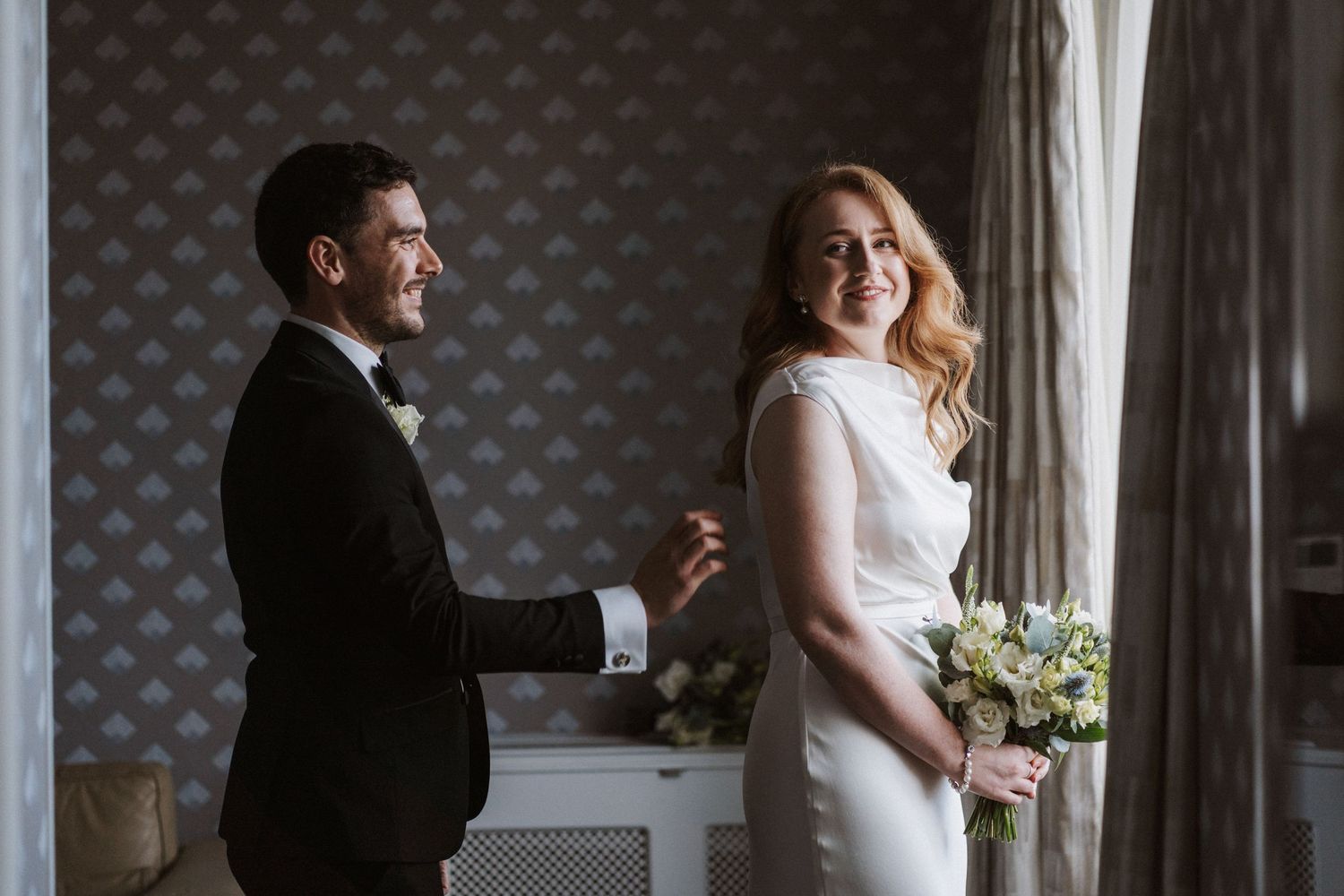 Couple shares intimate moment by window with elegant wallpaper and wedding attire.