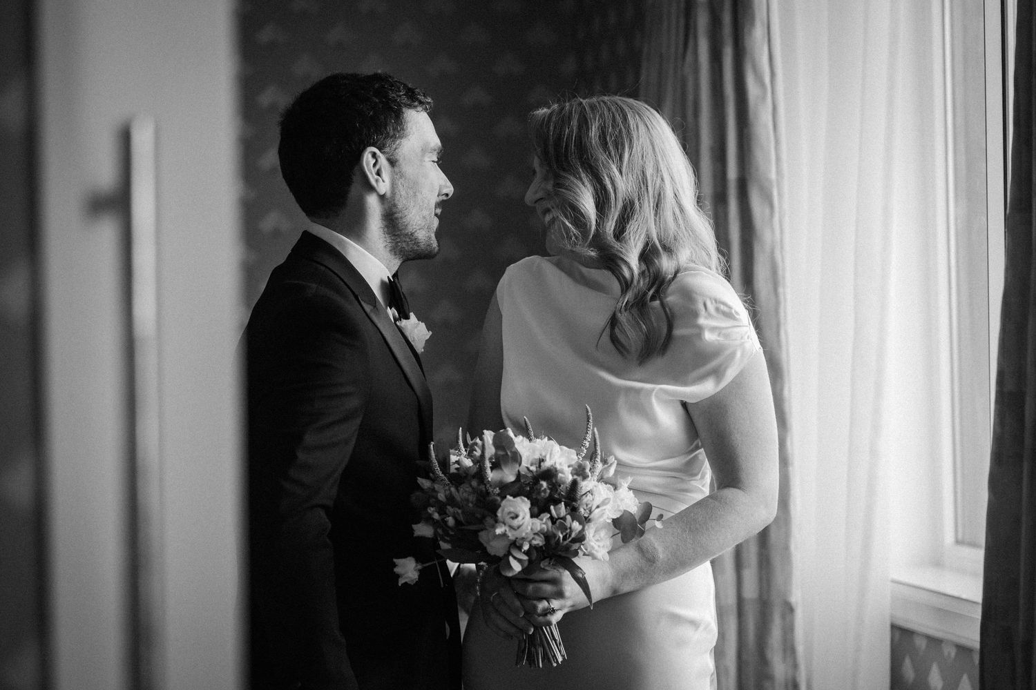 A couple shares an intimate moment during their wedding day in a softly lit room by a window.